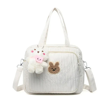Le Sac pour la maternité | longchamp est un sac à langer matelassé blanc avec des bretelles, une longue bandoulière amovible, une poche avant avec un visage de lapin marron et un porte-clés ours en peluche avec un nœud rose, idéal pour les parents en déplacement.